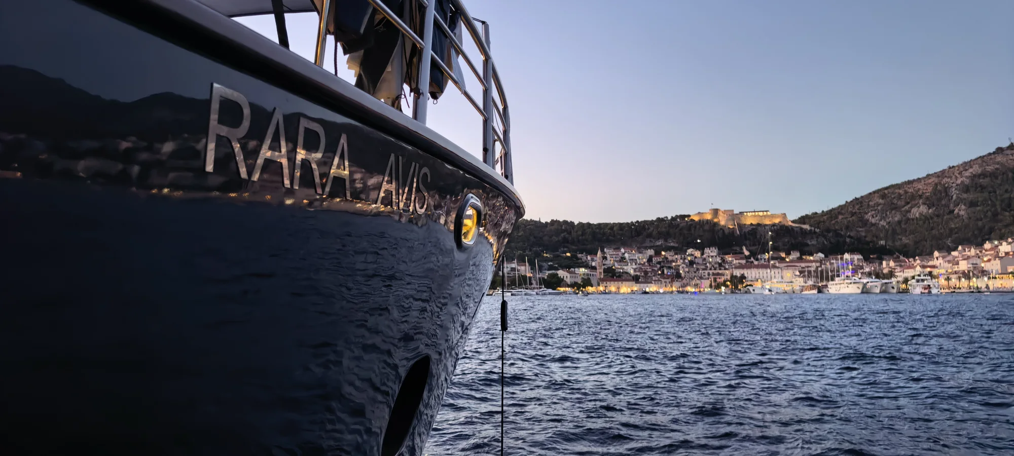 The bow of S/Y Rara Avis with Hvar Town and its fortress beyond, at dusk