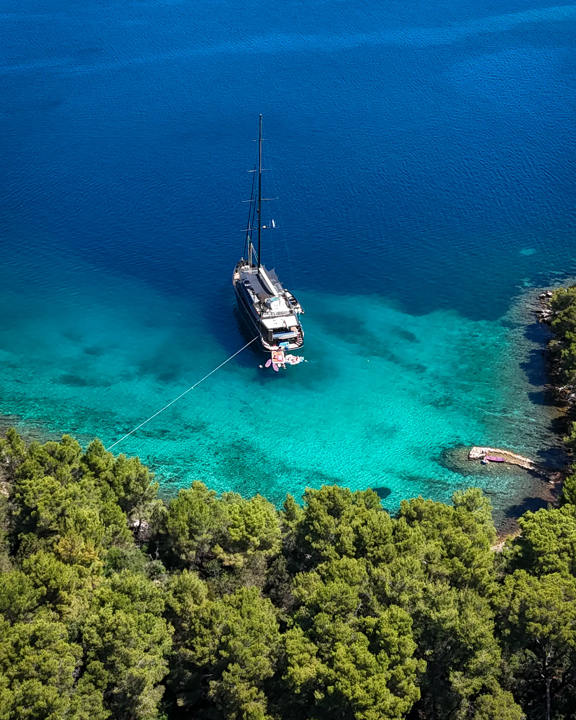 Aerial view of S/Y Rara Avis anchored in a pine-fringed Croatian bay, tender line run to shore