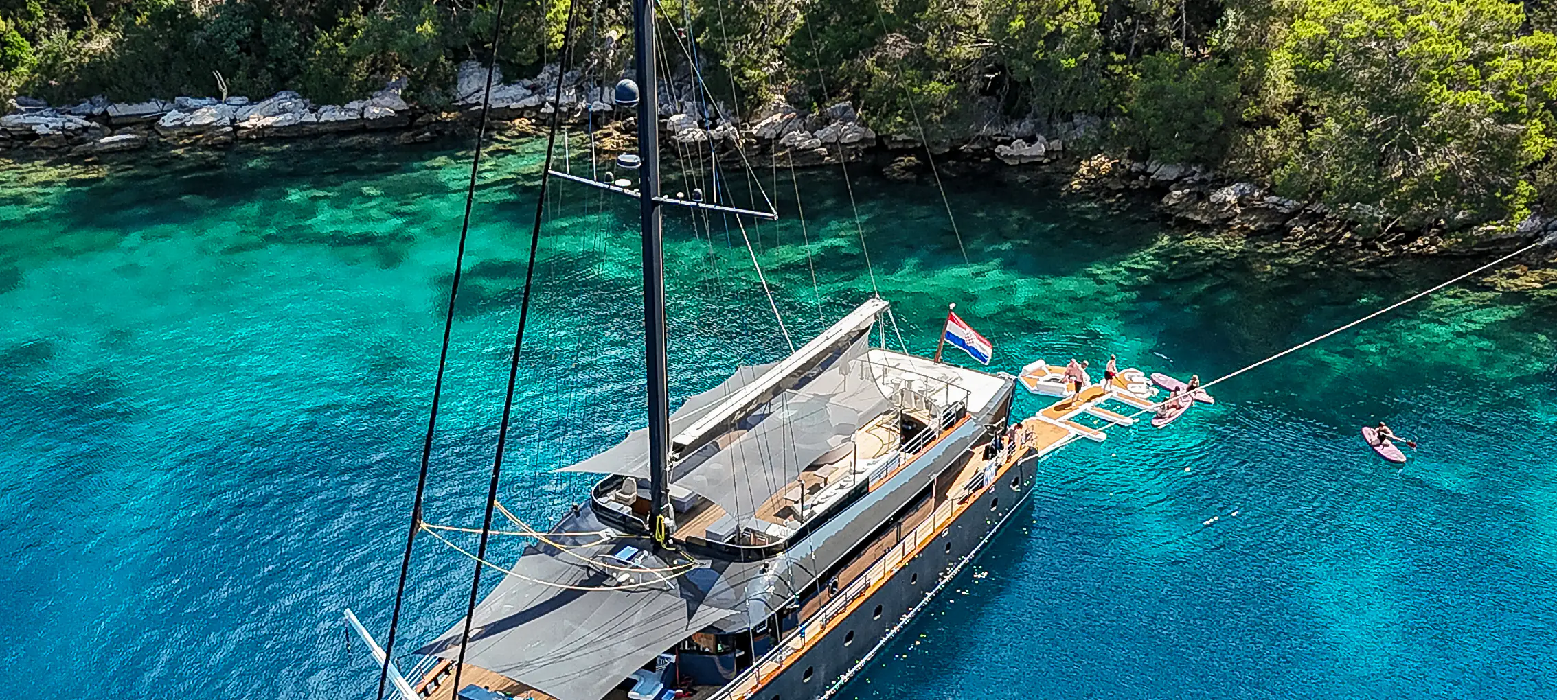 S/Y Rara Avis anchored in a pine-fringed Croatian cove, guests swimming from the floating platform